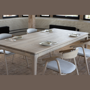 The Floating Dining Table seats six with plates, forks, and knives arranged at each spot. Light wood chairs with gray cushions surround it. The room has brick floors, cream walls, and black-framed windows.