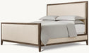 The Hampshire Bedframe features a dark wood frame with beige upholstered headboard and footboard, styled with light gray bedding and two stacked pillows, all set against a simple light background.