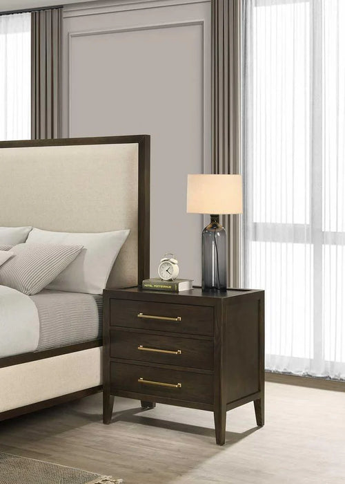 A modern bedroom features a beige upholstered bed beside the Hampshire Locker, a dark wooden nightstand with gold handles, topped with a gray lamp, a book, and a small décor piece. Light filters in through sheer curtains.