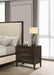 A modern bedroom features a beige upholstered bed beside the Hampshire Locker, a dark wooden nightstand with gold handles, topped with a gray lamp, a book, and a small décor piece. Light filters in through sheer curtains.