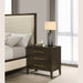 A modern bedroom features a beige upholstered bed beside the Hampshire Locker, a dark wooden nightstand with gold handles, topped with a gray lamp, a book, and a small décor piece. Light filters in through sheer curtains.