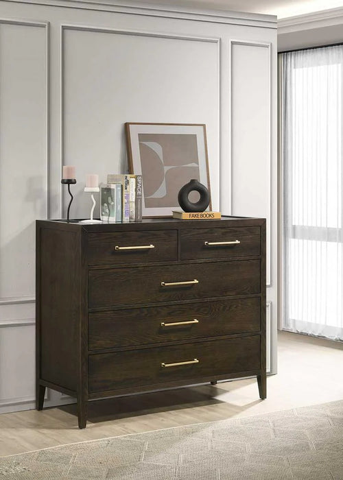 The Hampshire Tall Chest, featuring a dark wood finish and gold handles, stands against a paneled wall. On top are candles, books, abstract art, and a black ornament, while sunlight filters through a sheer curtain on the right.