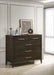The Hampshire Tall Chest, featuring a dark wood finish and gold handles, stands against a paneled wall. On top are candles, books, abstract art, and a black ornament, while sunlight filters through a sheer curtain on the right.