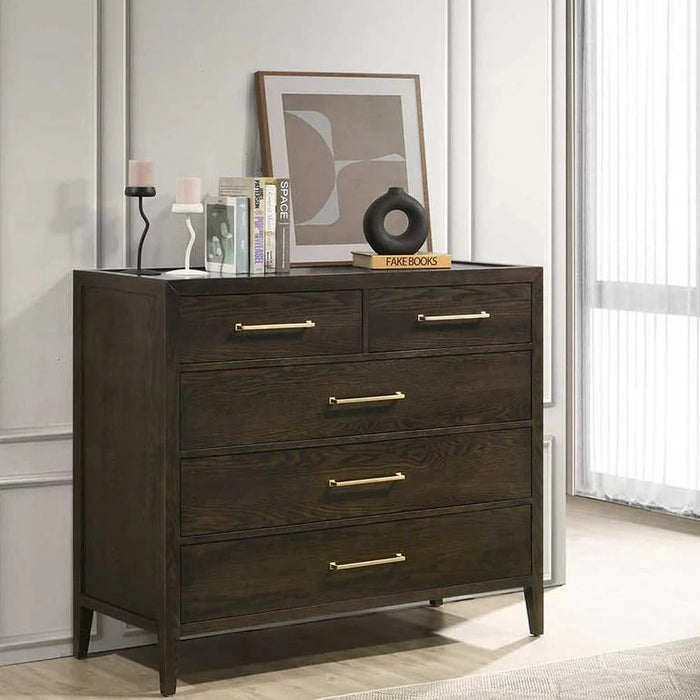 The Hampshire Tall Chest, featuring a dark wood finish and gold handles, stands against a paneled wall. On top are candles, books, abstract art, and a black ornament, while sunlight filters through a sheer curtain on the right.