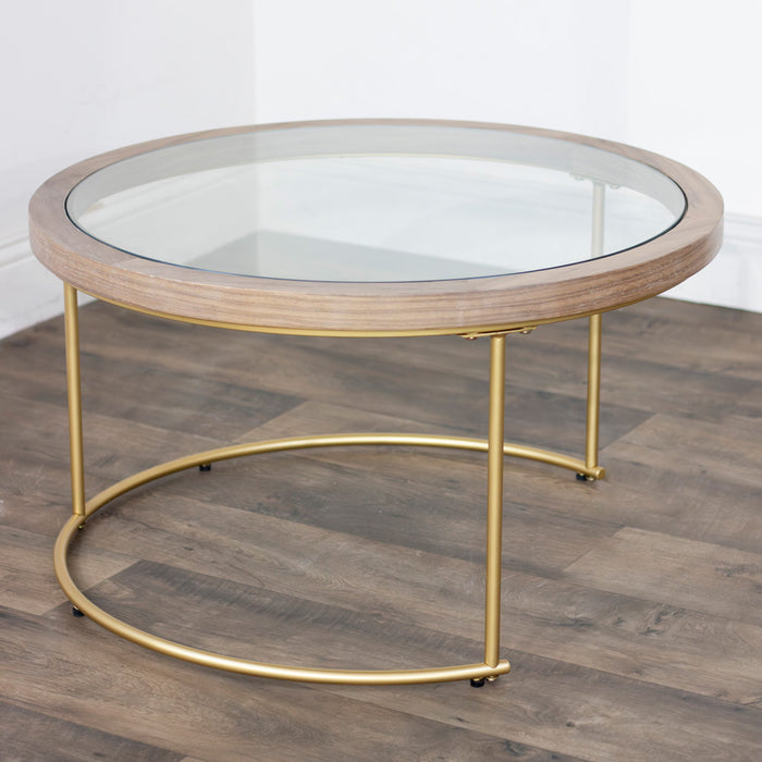 The Hannah Coffee Table features wood accents, a glass top, and elegant gold legs, shown on a wooden floor near a white wall in a stylish room.