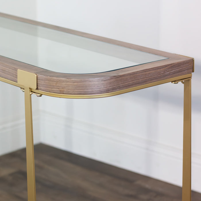 A close-up of the Hannah Console Table, featuring striking wood details, a glass top, and gold legs, set on a dark wood floor against a white wall.