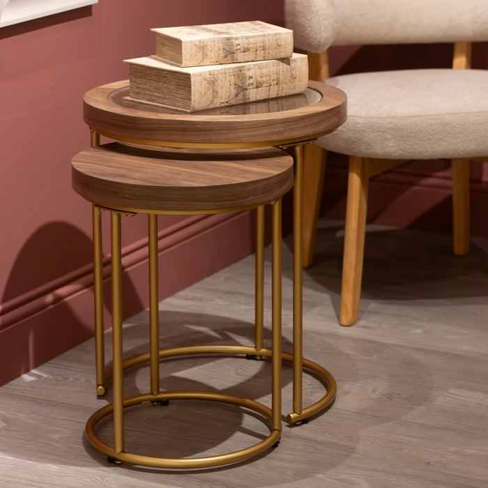 A pair of round Hannah Nest of Tables with wood tops and modern gold legs sits on a wood floor by a beige chair. The larger table holds a stack of three books with light brown covers.