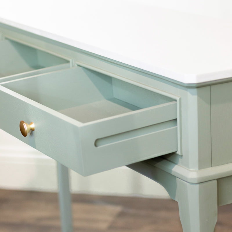 A close-up of the Monroe Large Console Table with a white top, showing an open drawer with a round gold knob. The sage table stands on four legs and is placed on a wood floor.