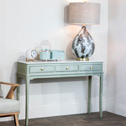 The Monroe Large Console Table with gold knobs holds a marble-patterned lamp, a clock, a diffuser, and a small mint green box. This sage 3-drawer console stands against a white wall next to a light wood and gray armchair.