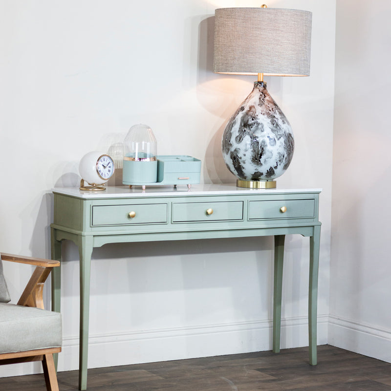 The Monroe Large Console Table with gold knobs holds a marble-patterned lamp, a clock, a diffuser, and a small mint green box. This sage 3-drawer console stands against a white wall next to a light wood and gray armchair.