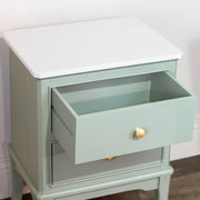 The Monroe Locker is a sage 2-drawer accent table with round gold knobs and a white marble-patterned top. The upper drawer is partially open, displaying its elegant design against a white wall and wooden floor.