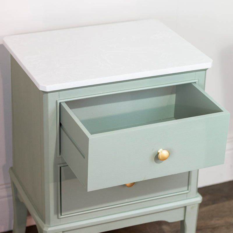 The Monroe Locker is a sage 2-drawer accent table with round gold knobs and a white marble-patterned top. The upper drawer is partially open, displaying its elegant design against a white wall and wooden floor.