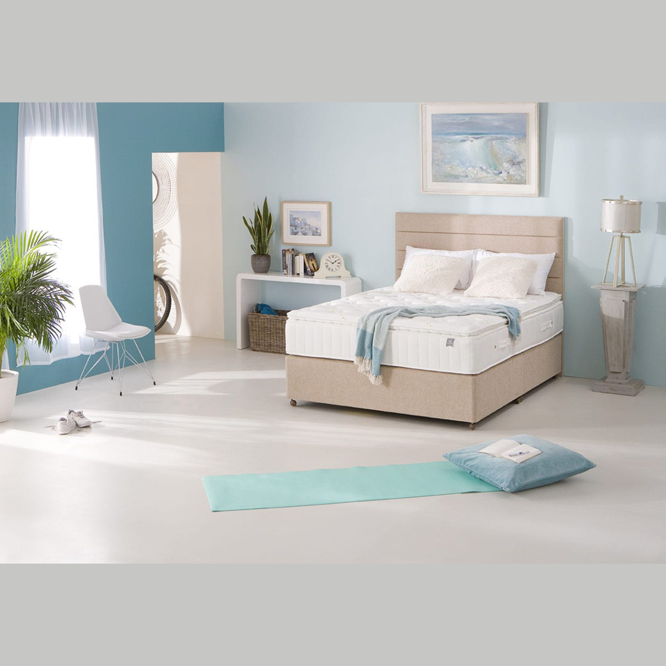 A bright, minimalist bedroom with a beige bed featuring the King Koil - Spinal Pocket Pillow Top mattress, white bedding, and a pastel blue accent wall. A yoga mat and pillow rest on the floor as sunlight streams through sheer curtains.